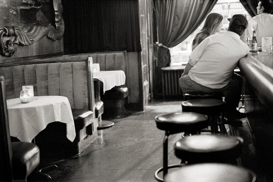 A man and a woman sit at the bar, talking. Daylight streams in the front window.