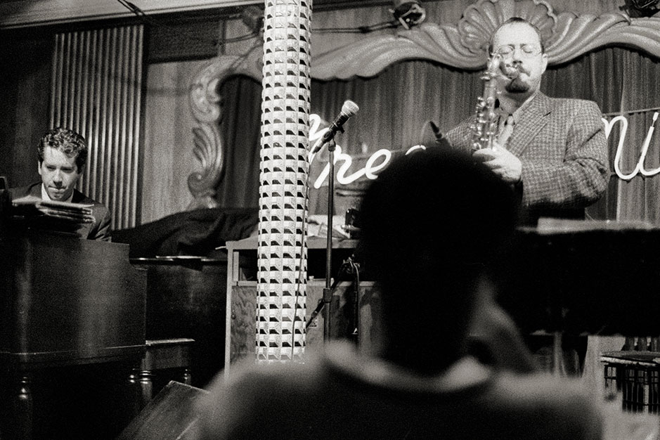A man listens to a saxophonist taking a solo. The piano player, visible at the left edge of the image, listens too.