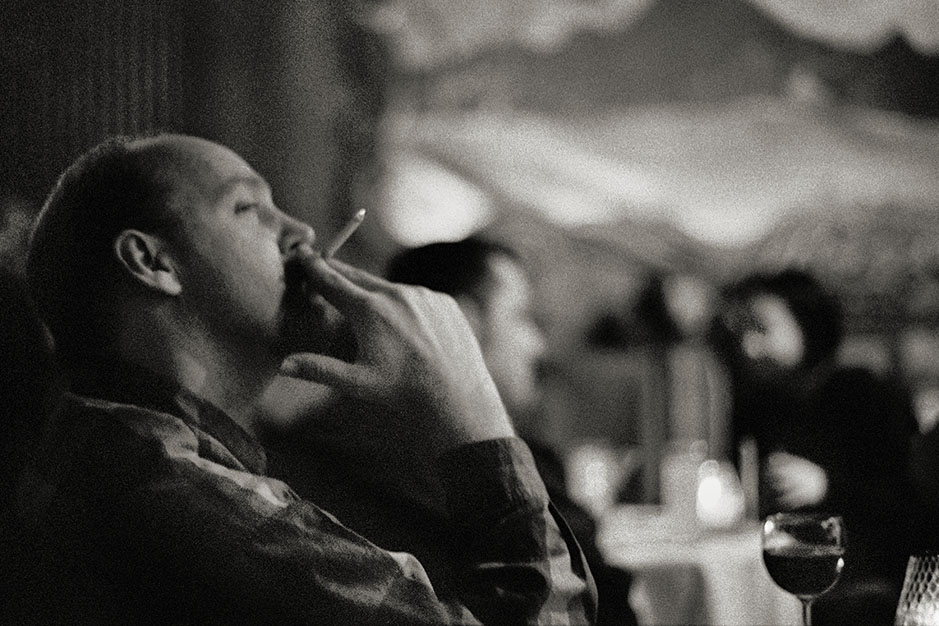 A man drinks a glass of wine and smokes a cigarette, facing the stage.