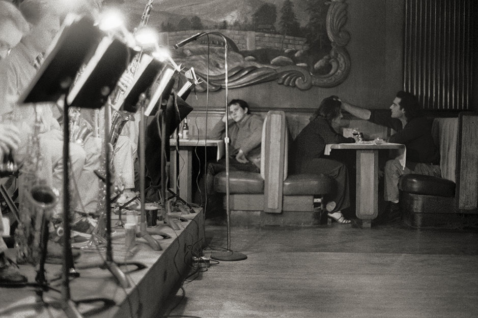 A man listens to the band. At a nearby table, a man places his hand on a woman's head.