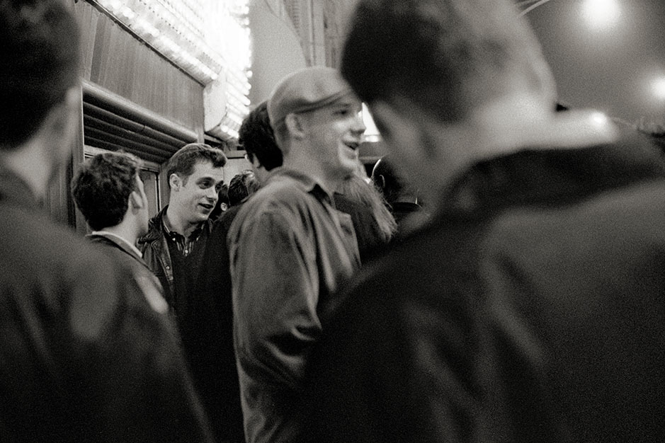 A young man wearing a leather jacket stands in line outside, talking to his friends.