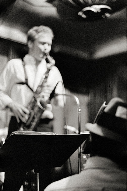 A saxophonist plays a solo while a man in the audience sits listening.